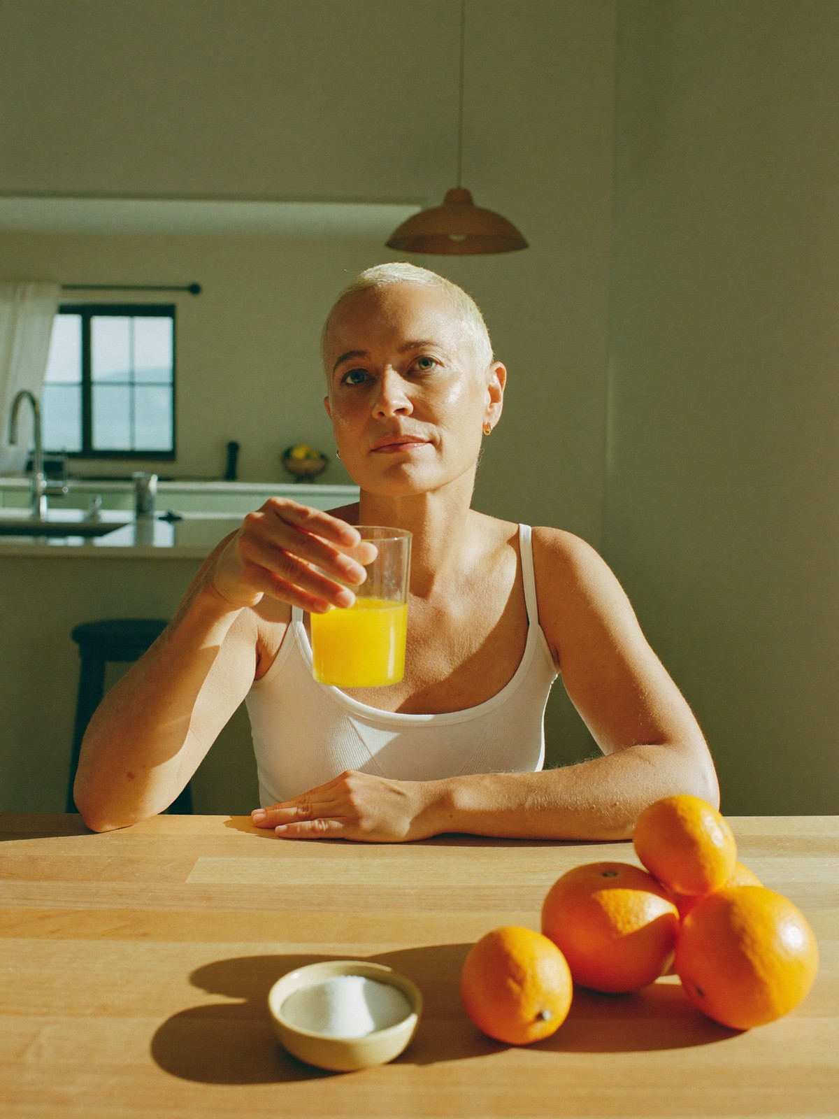 A woman over 40 holding a glass of orange citrus drink at a sunlit kitchen table with fresh oranges