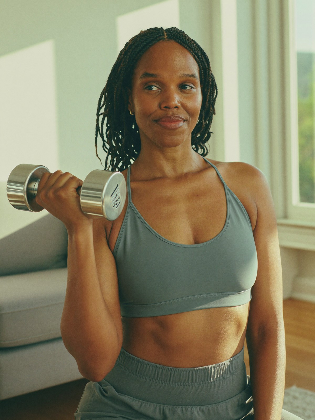 A woman over 40 doing a light dumbbell curl in a bright, sunlit living room